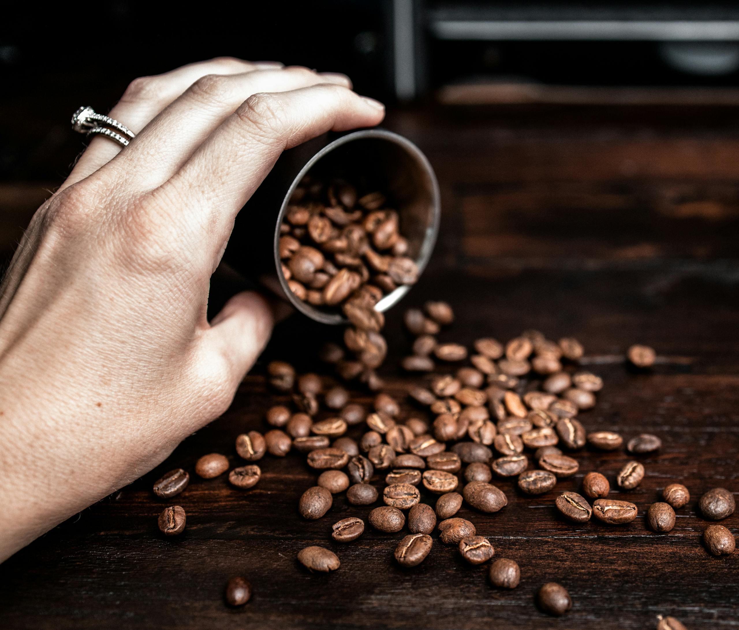 A hand pours aromatic coffee beans from a container onto a wooden surface, highlighting rich flavor and aroma.