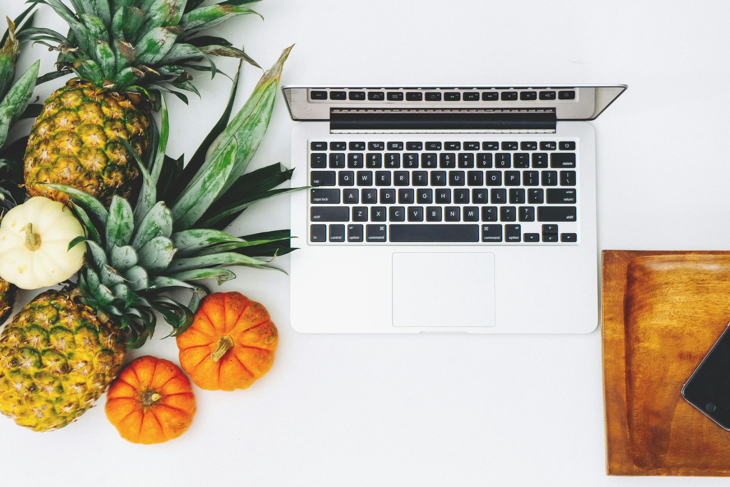 Stylish workspace featuring a laptop, pineapples, and pumpkins on a white background.
